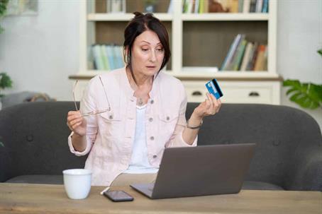 A woman is sitting on the couch and looking at her laptop in disbelief as she holds her glasses and a blue credit card in her hands.